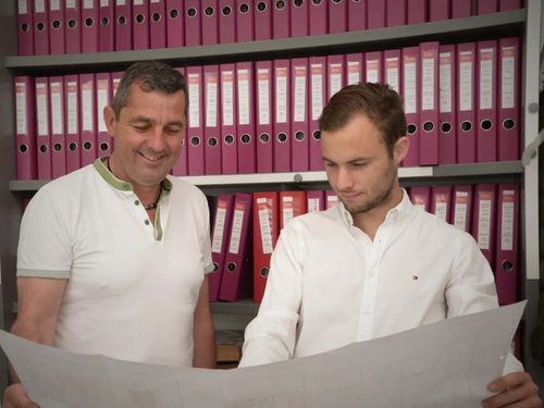 Zwei Männer in weißen Hemden stehen und schauen sich ein Dokument an, mit Regalen voller pinker und roter Ordner dahinter.