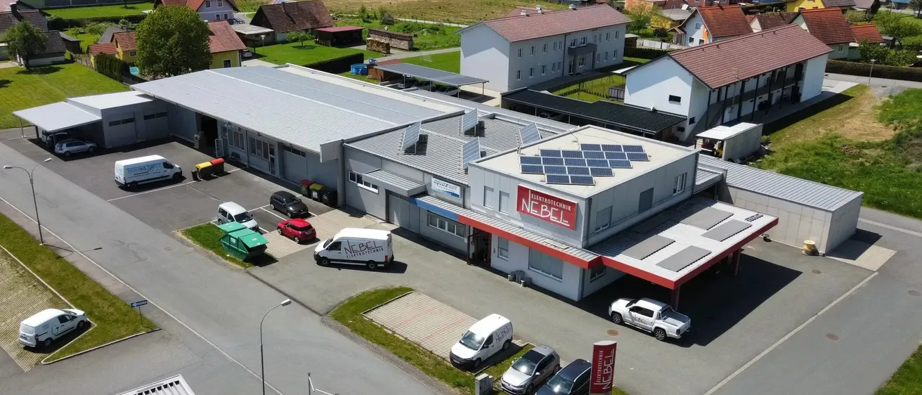 Eine Luftaufnahme des Gebäudes von NEBEL Elektrotechnik mit Solarpanelen auf dem Dach, mehreren geparkten Fahrzeugen davor und einem geparkten Pickup-Truck in der Ferne.