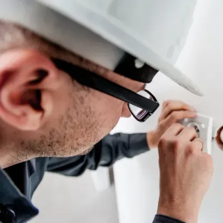 Ein Mann in einem weißen Schutzhelm und Brille repariert eine Steckdose an einer weißen Wand.