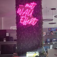 Ein Neonschild mit den Worten 'mit WAU Effekt' ist auf einer vertikalen lila Pflanzenwand prominent angebracht. Darunter ist ein 'HONOR 20' Schild zu sehen, und Regale mit verschiedenen Produkten sind sichtbar.