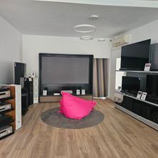 Ein modernes Wohnzimmer mit einem großen Flachbildfernseher und einem rosa Pouf auf einem runden Teppich. Mehrere Lautsprecher und elektronische Geräte sind auf Regalen platziert.