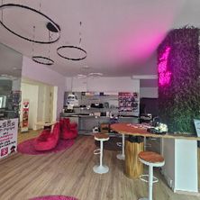 Ein Innenraumdesign mit einer rosa Wand, einem Holzboden und einem rosa Teppich. Es gibt runde Hocker und einen Holztisch mit einem rosa Neonschild. Es gibt eine Theke mit einer Glastür und eine große Pflanze auf der rechten Seite.