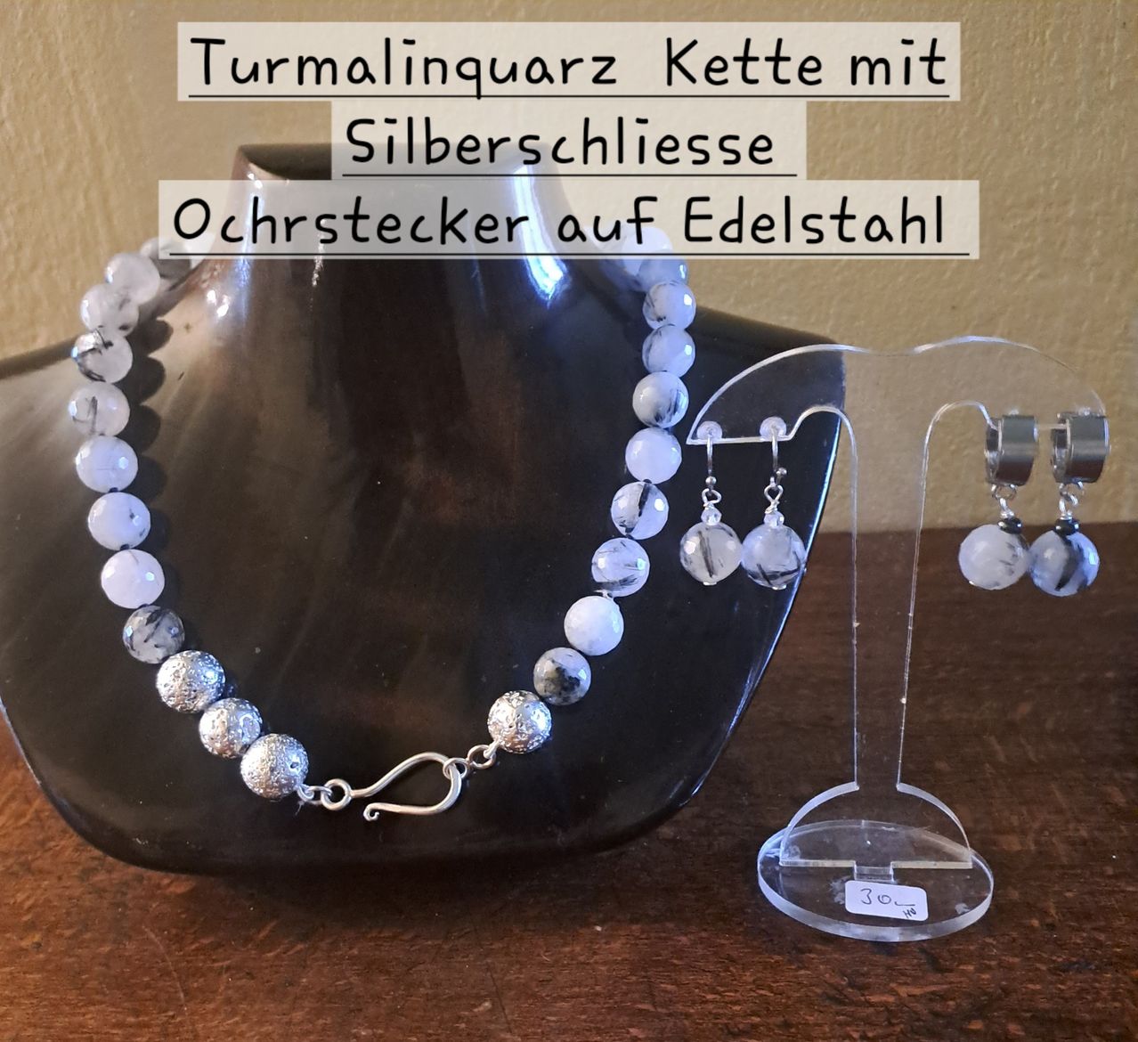 A jewelry set with a white and gray beaded necklace and earrings displayed on a wooden surface with a glass stand.