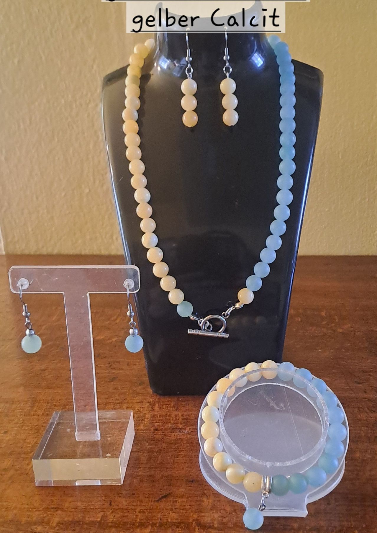 A beaded necklace with white and blue beads, along with matching earrings, rests on a black display. Below, a bracelet with similar beads is on a wooden surface.
