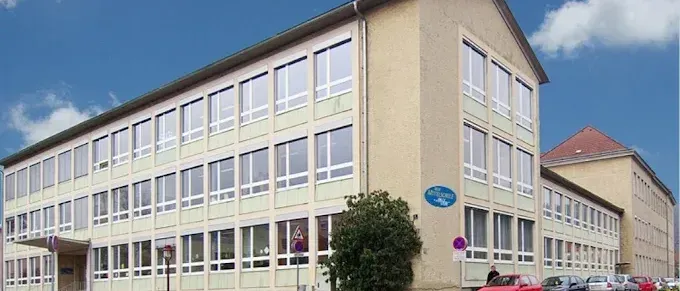 Ein großes Bürogebäude mit vielen Fenstern steht unter einem teilweise bewölkten Himmel. Ein rotes Auto und andere Autos sind vor dem Gebäude geparkt. Eine Person steht in der Nähe eines Schildes.