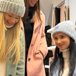 Zwei lächelnde Frauen in einem Geschäft, eine in einer grünen Jacke und die andere in einer rosa Jacke, beide tragen Mützen. Hinter ihnen hängen Jacken an einem Kleiderständer.
