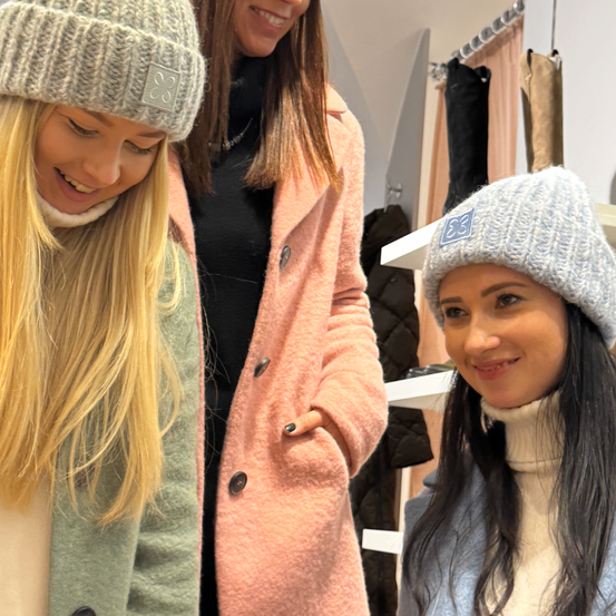 Zwei lächelnde Frauen in einem Geschäft, eine in einer grünen Jacke und die andere in einer rosa Jacke, beide tragen Mützen. Hinter ihnen hängen Jacken an einem Kleiderständer.