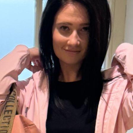 Eine Frau mit langen schwarzen Haaren lächelt und posiert für ein Foto. Sie trägt eine rosa Jacke und ein schwarzes Oberteil. Sie hält ihre Jacke mit der linken Hand.