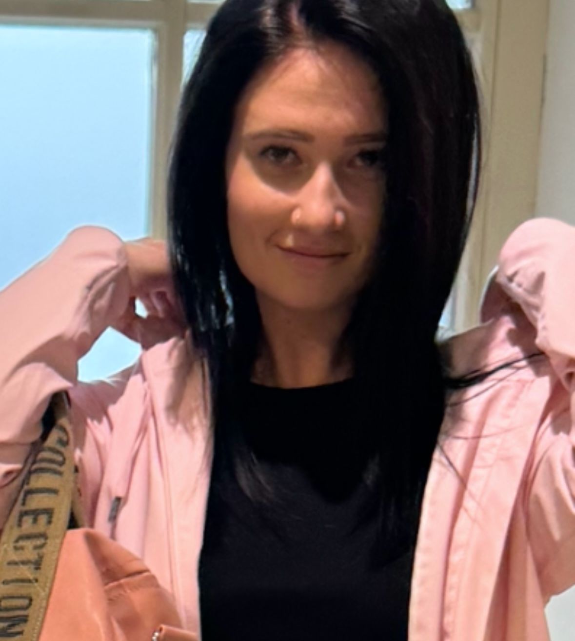 Eine Frau mit langen schwarzen Haaren lächelt und posiert für ein Foto. Sie trägt eine rosa Jacke und ein schwarzes Oberteil. Sie hält ihre Jacke mit der linken Hand.