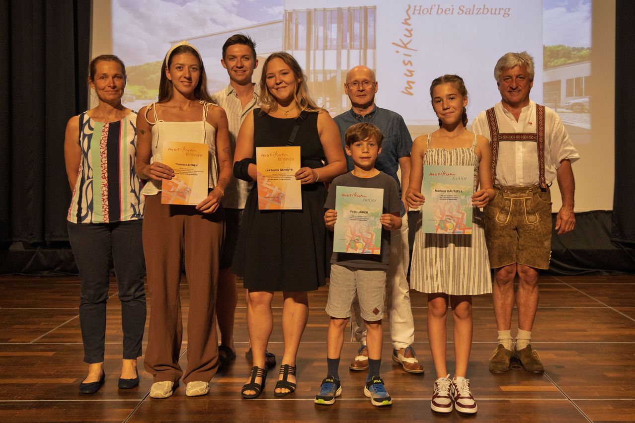 Eine Gruppe von Menschen posiert für ein Foto mit Urkunden in den Händen. Sie stehen auf einer Bühne, mit einer Projektionsleinwand hinter ihnen. Der Junge in der Mitte hält eine Urkunde mit einem farbenfrohen Design. Die beiden Frauen auf beiden Seiten von ihm lächeln und schauen in die Kamera.