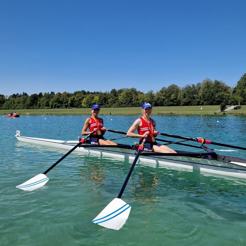 Zwei Frauen in roten Rettungswesten und blauen Mützen rudern ein Boot auf einem ruhigen See mit Bäumen im Hintergrund.