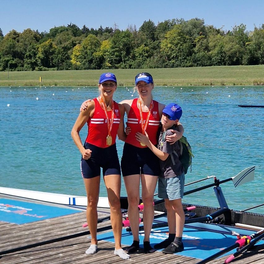 Drei Frauen und ein Junge stehen auf einem Steg am See, tragen Medaillen und lächeln. Die Frauen tragen rote Oberteile und schwarze Shorts, der Junge trägt einen blauen Hut.
