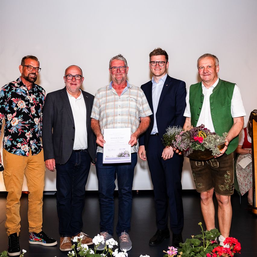 Fünf Männer stehen auf einer Bühne, einer hält ein Zertifikat und ein anderer eine Blumenstrauß. Sie sind in verschiedenen Outfits gekleidet, und eine Harfe ist im Hintergrund zu sehen.
