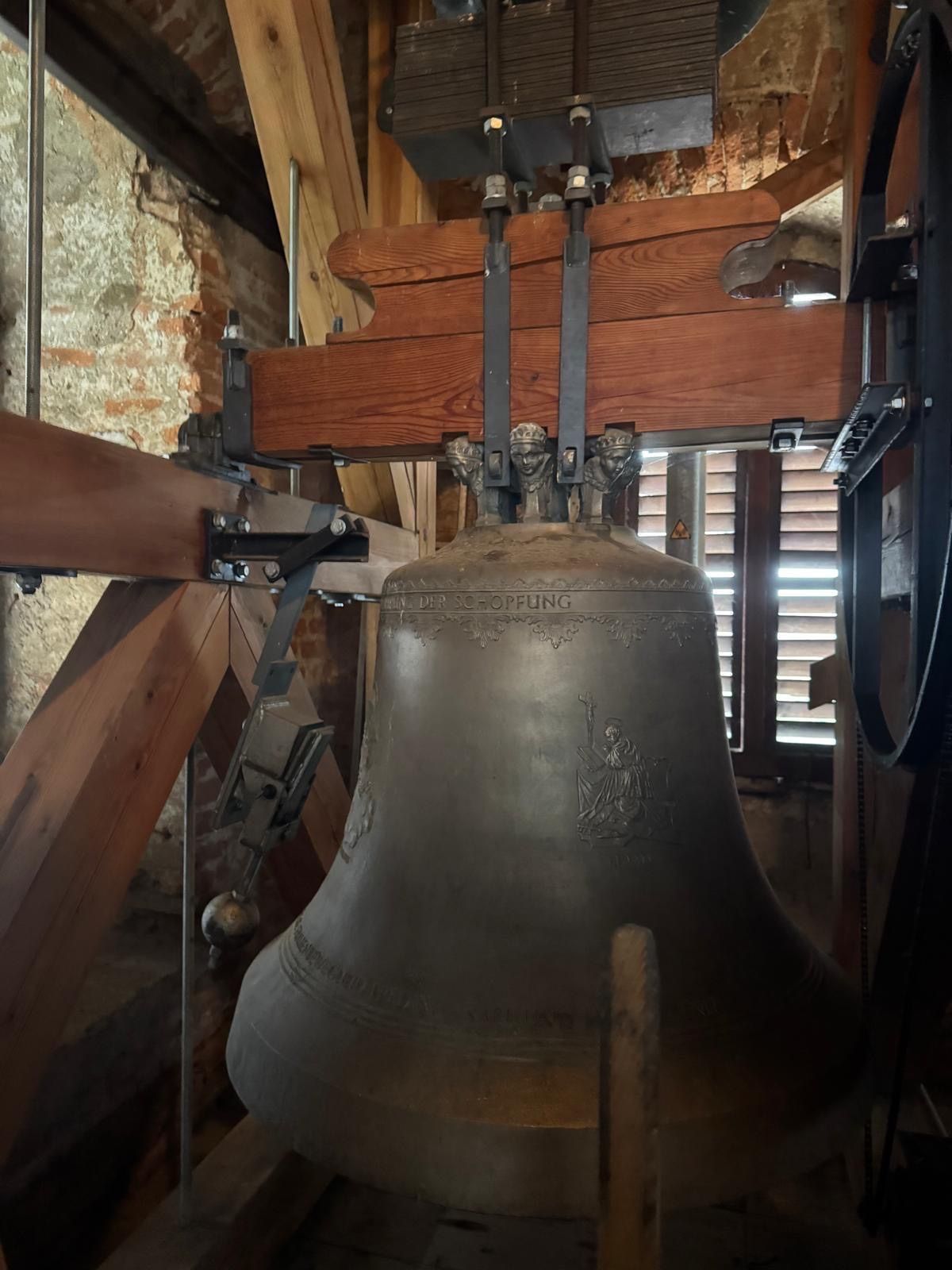 Eine große, alte Glocke mit kunstvollen Schnitzereien hängt von einem Holzbalken in einem Glockenturm. Die Glocke ist mit 'Der Schopfling' beschriftet und zeigt eine Frauenfigur.