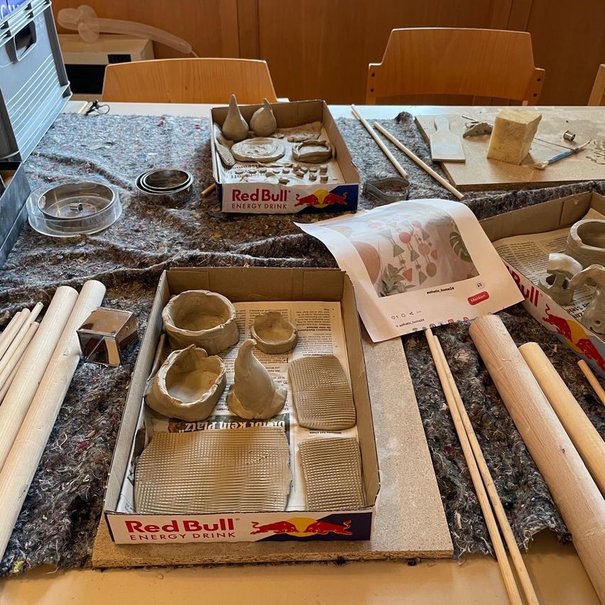 Ein Arbeitsbereich mit Tonfiguren, Holzstäben und einer Red Bull-Box auf einem mit grauem Stoff bedeckten Tisch.