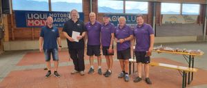 Fünf Männer in lila Hemden stehen zusammen und halten Gegenstände. Ein Mann hält ein Papier, und ein anderer hat eine Geldbörse. Hinter ihnen zeigt eine Holzwand Banner, und eine Bank ist rechts platziert.