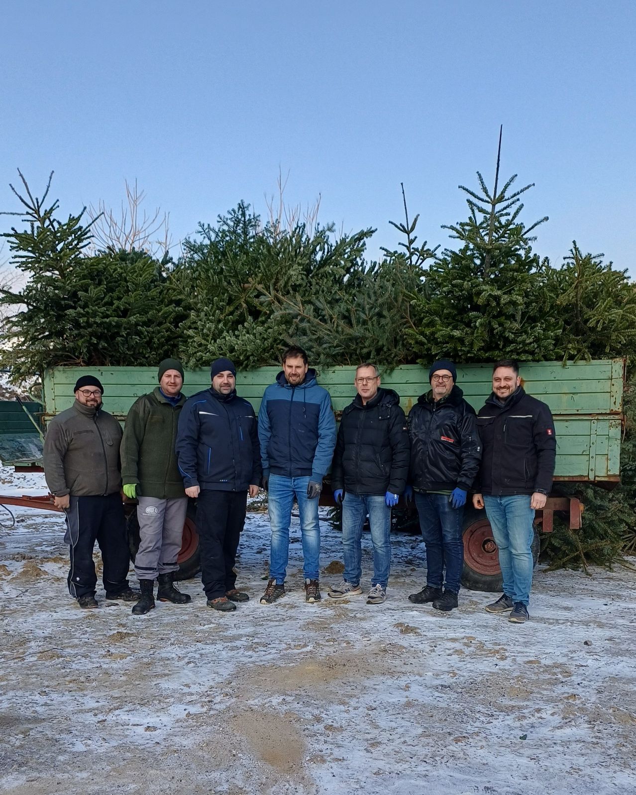 A group of men are standing in front of a trailer loaded with pine trees, wearing winter clothing and gloves.