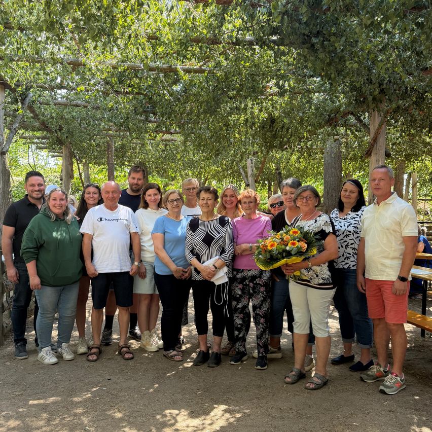 Gruppe von Menschen steht unter Bäumen, eine Frau hält Blumenstrauß