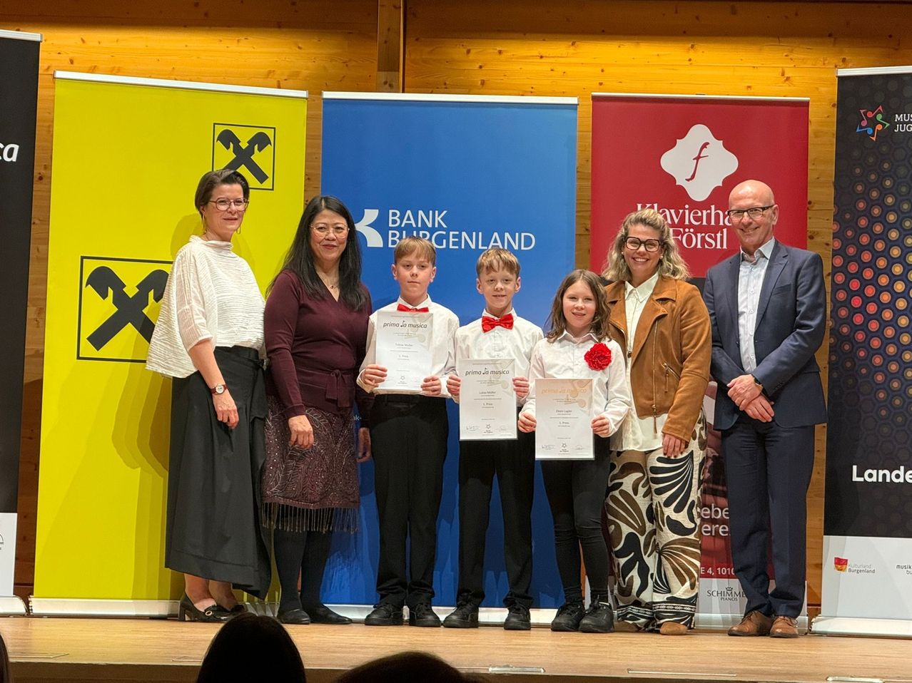 Drei Kinder mit Zertifikaten stehen auf einer Bühne mit Erwachsenen. Sie alle lächeln. Hinter ihnen sind Banner mit Logos und Texten. Ein Banner hat den Text 'Bank Burgenland'.