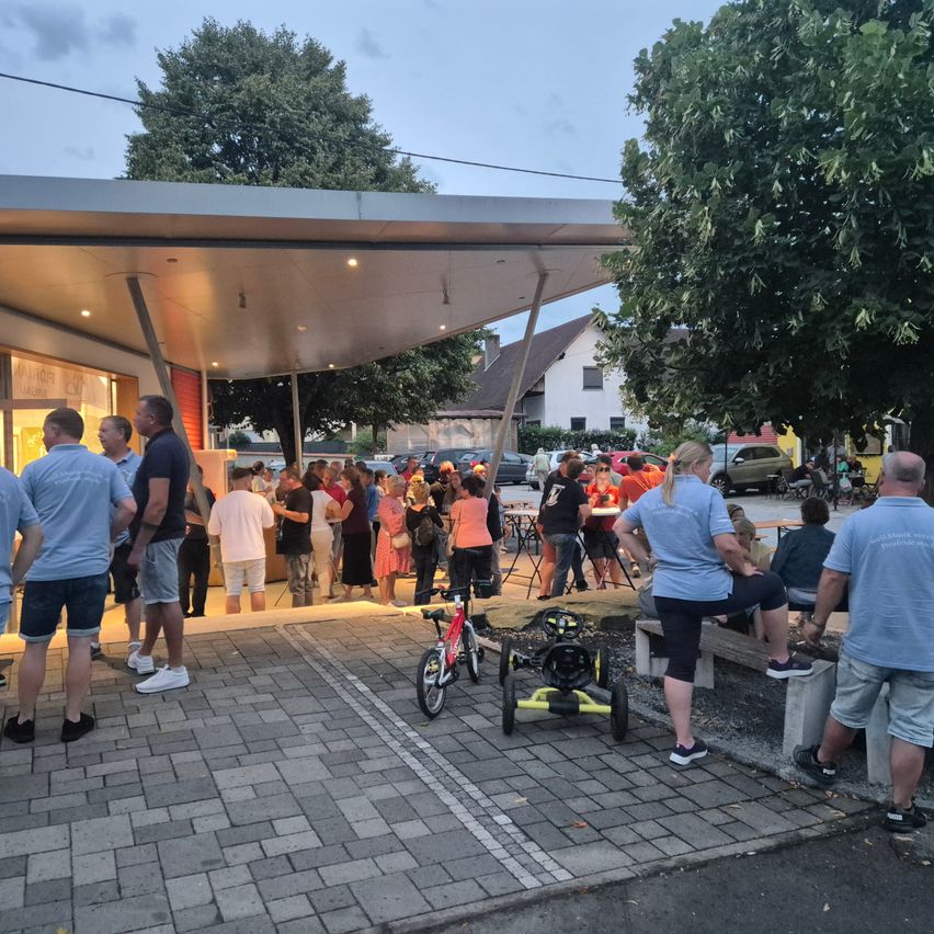 Eine Gruppe von Menschen versammelt sich vor einem Imbisswagen, einige stehen in der Schlange und andere sitzen auf Bänken. In der Nähe sind Fahrräder geparkt, und ein Baum spendet Schatten. Im Hintergrund befinden sich Häuser und geparkte Autos.