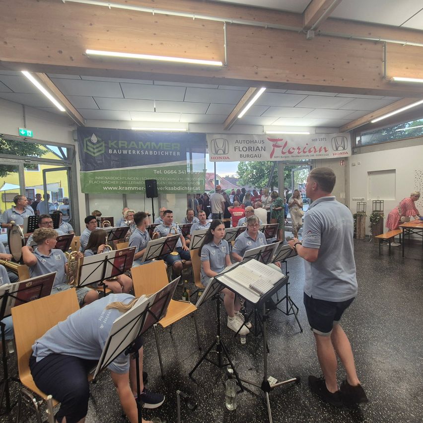 Ein Orchester probt in einem Raum mit Holzstühlen und Notenständern. Ein Mann steht vor ihnen, möglicherweise ihr Dirigent. Es hängen Banner an der Wand.