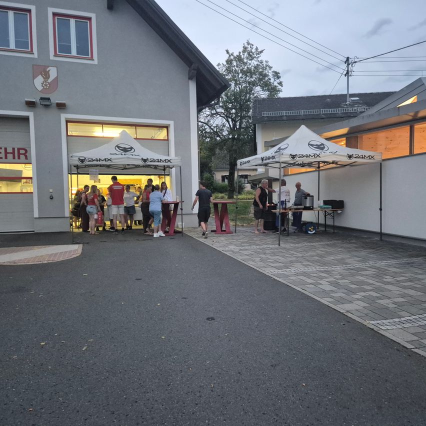 Eine Gruppe von Menschen steht vor einem Gebäude mit zwei aufgestellten Zelten. Ein Zelt hat einen roten Tisch, und das andere hat einen Tisch mit einer schwarzen Tasche. Das Gebäude hat ein Logo an der Wand.