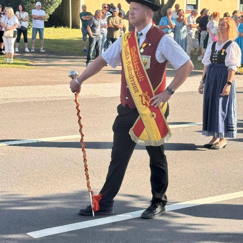 Ein Mann in traditioneller Kleidung geht in einem Umzug, hält einen zeremoniellen Stab und trägt eine Schärpe. Zuschauer stehen hinter ihm an der Straße.