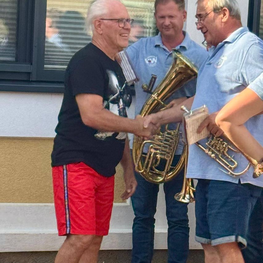 Drei Männer stehen draußen, einer schüttelt die Hand eines anderen und hält Blechblasinstrumente. Der Mann in Schwarz schüttelt die Hand des Mannes in Blau.