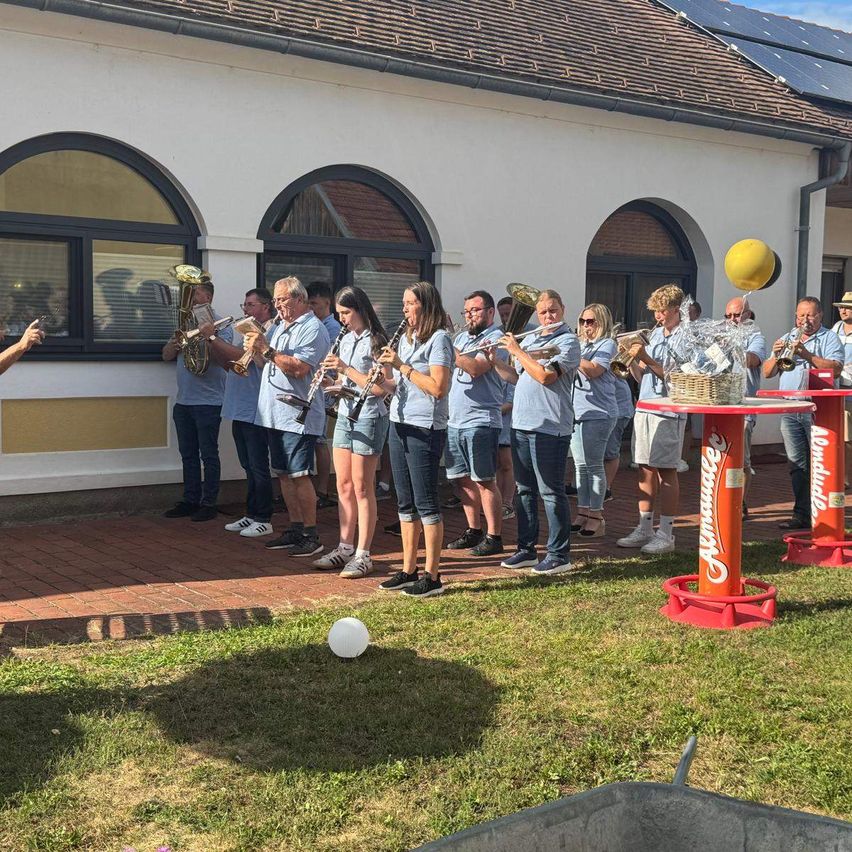Eine Gruppe von Musikern in blauen Shirts und Shorts steht vor einem Gebäude mit Rundbogenfenstern und spielt verschiedene Instrumente. Eine Person hält ein Telefon vor ihnen.