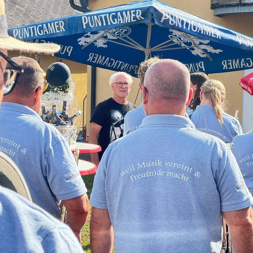 Eine Gruppe von Menschen in blauen Shirts mit dem Text 'Weil Musik vereint & Freud(n)de macht' steht unter einem blauen Schirm mit dem Text 'Puntigamer'. Ein Mann in einem schwarzen Shirt steht hinter ihnen.