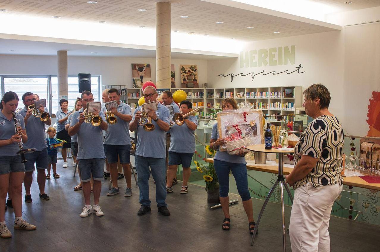 Eine Gruppe von Männern in blauen Hemden und Shorts spielt Trompeten in einer Bibliothek, während eine Frau in blauen Jeans einen Korb mit Geschenken hält.