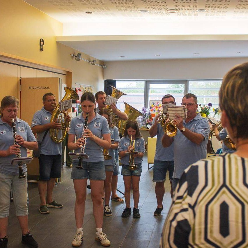 Eine Gruppe von Musikern in blauen Shirts spielt verschiedene Instrumente in einem Raum mit gelben Wänden. Eine Frau beobachtet sie.