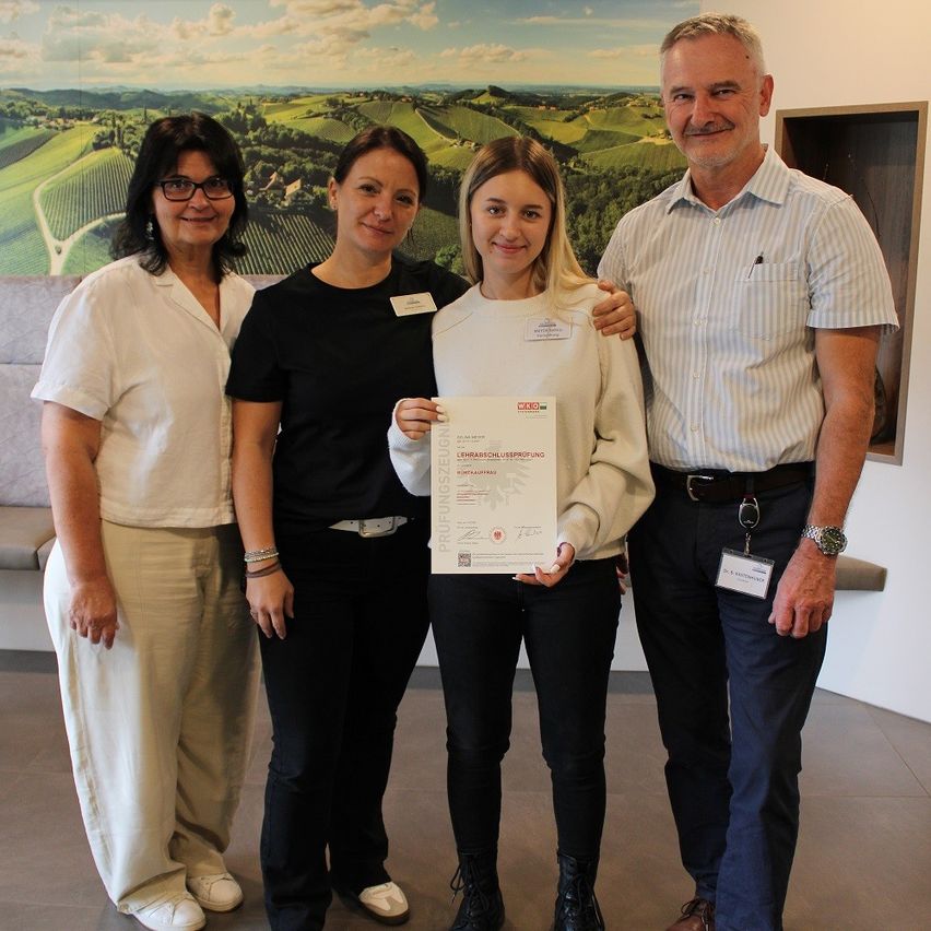 Eine junge Frau hält ein Zertifikat in der Hand, während sie zwischen zwei Frauen und einem Mann steht, alle lächeln und posieren für ein Foto in einem Raum mit einer malerischen Kulisse.
