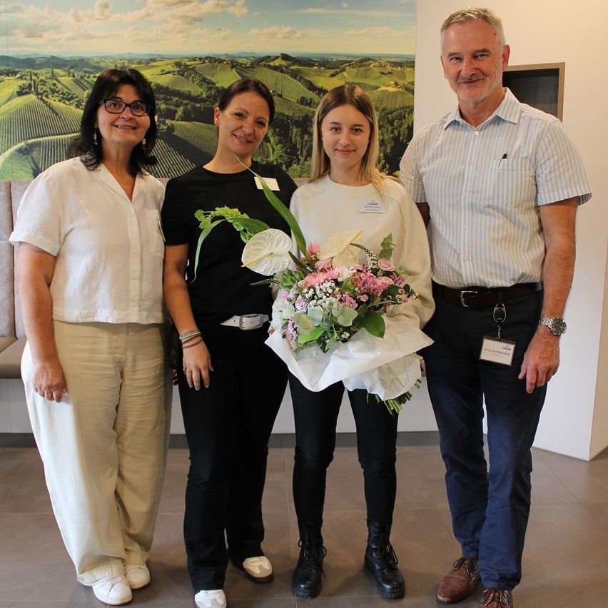Vier Erwachsene stehen in einem Raum und lächeln. Eine Frau hält einen Blumenstrauß. Hinter ihnen befindet sich ein großes Landschaftsgemälde.