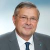 Ein Mann in einem grauen Anzug und blauer Krawatte mit Brille lächelt in die Kamera.