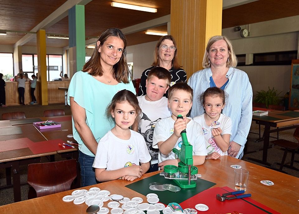 Vier Erwachsene und vier Kinder stehen gemeinsam in einem Klassenzimmer. Drei der Kinder posieren für ein Foto, während sie eine grüne Stanzmaschine halten.