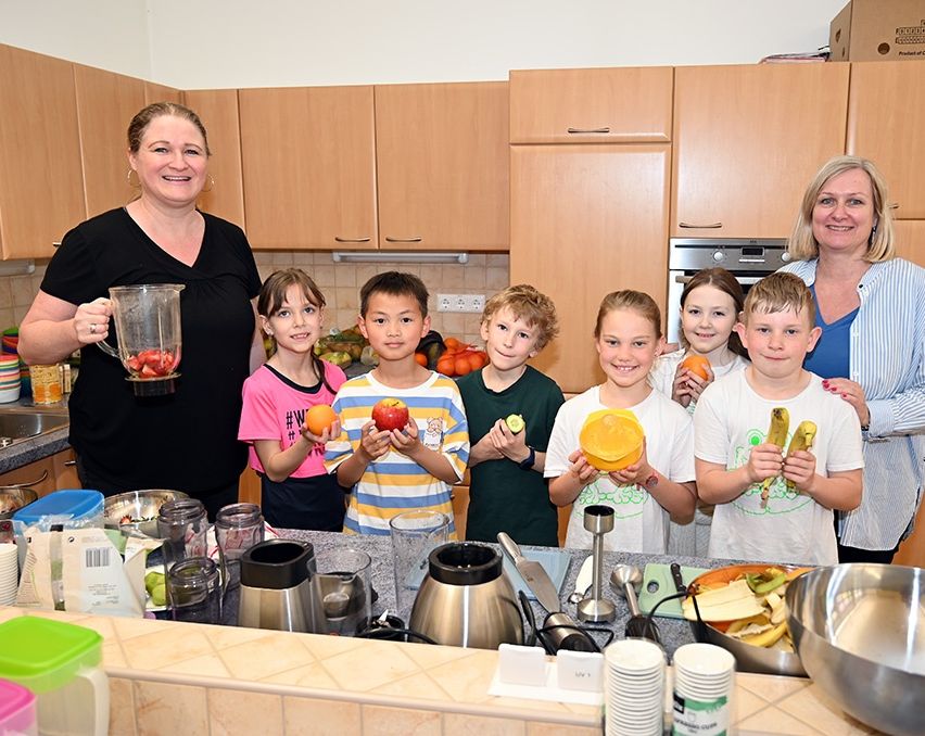 Eine Frau mit einem Mixer in der Küche, umgeben von lächelnden Kindern, die Obst halten. Hinter ihr steht eine weitere Frau mit einem Lächeln. Die Küche ist mit Schränken, Geräten und einer Arbeitsplatte mit Obst und Küchenutensilien ausgestattet.