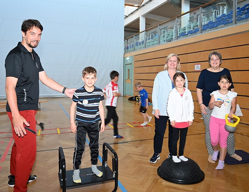 Ein Mann in einem schwarzen Shirt und roten Hosen steht neben einem jungen Jungen, der auf einer Plattform in einer Sporthalle balanciert. Dahinter stehen zwei Frauen und zwei Mädchen auf einem Balanceball.