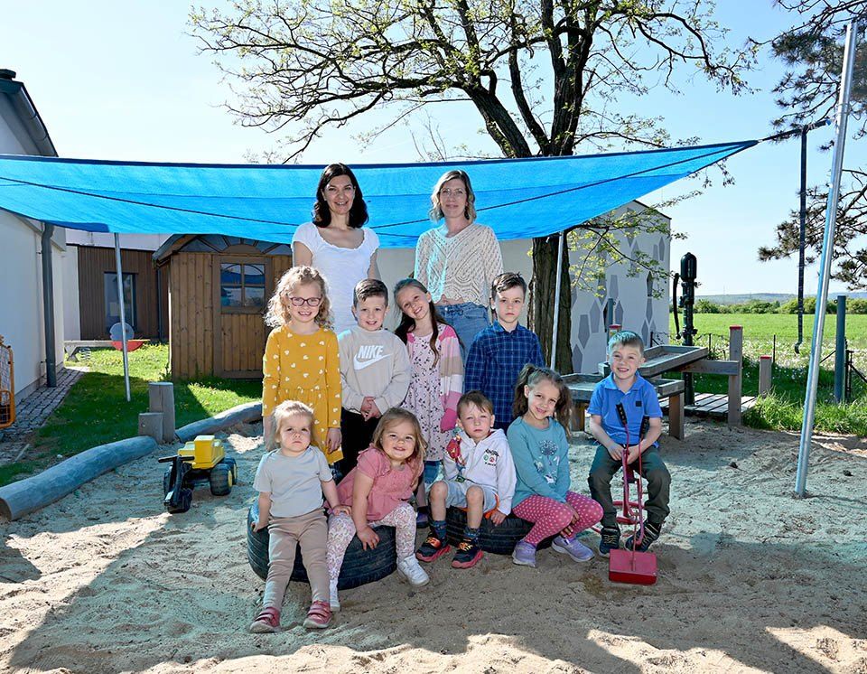 Eine Gruppe von Kindern und zwei Frauen sind in einem Spielplatz mit einem blauen Baldachin und Sandkasten aus Reifen. Dahinter sind ein Gebäude und ein Grasbereich zu sehen.