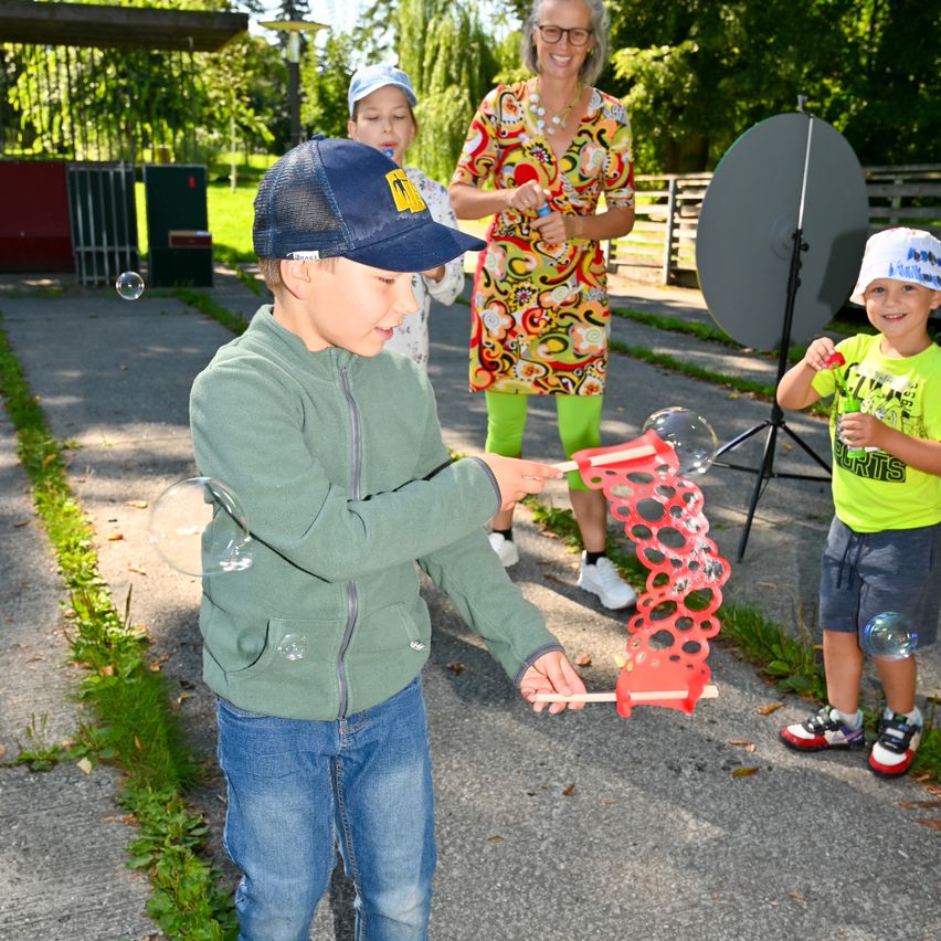 Zwei Jungen und eine Frau sind draußen, einer der Jungen bläst mit einem Blasebalg Seifenblasen. In der Nähe steht eine Satellitenschüssel. Bäume und ein Zaun sind im Hintergrund zu sehen.