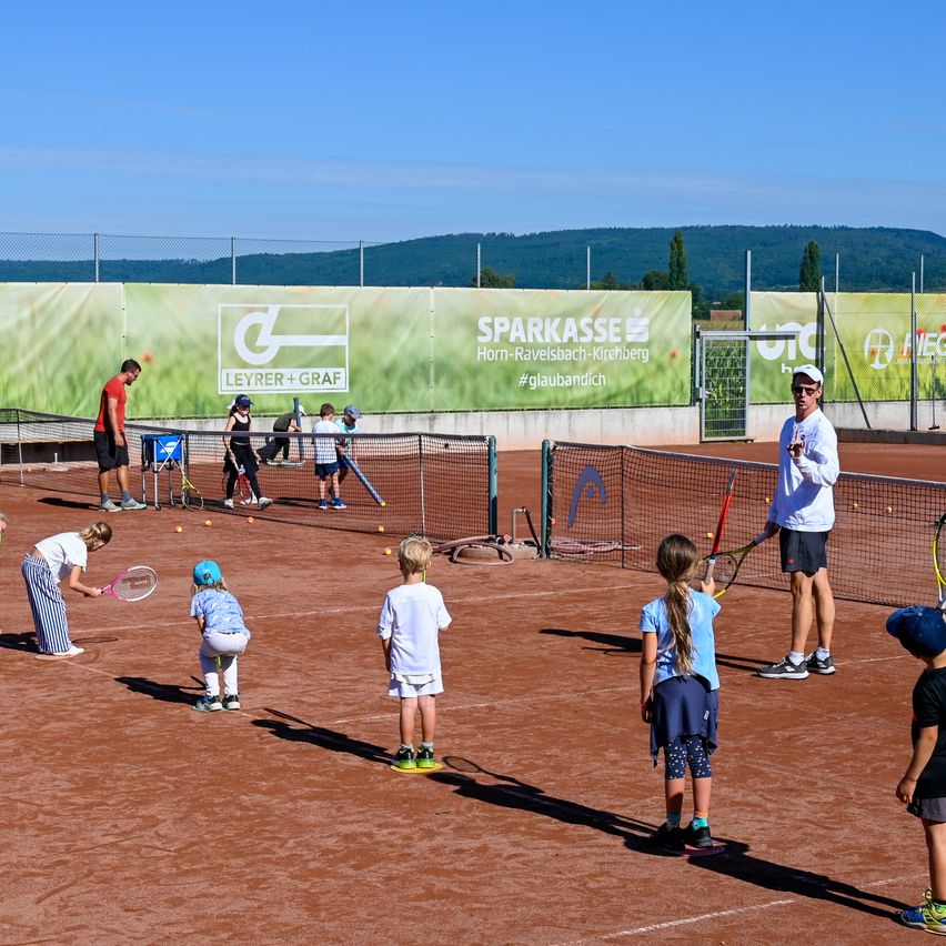 Eine Gruppe von Kindern lernt auf einem Tennisplatz Tennis zu spielen, wobei ein Mann sie anleitet. Einige halten Tennisschläger. Der Platz hat einen Zaun mit Bannern dahinter.