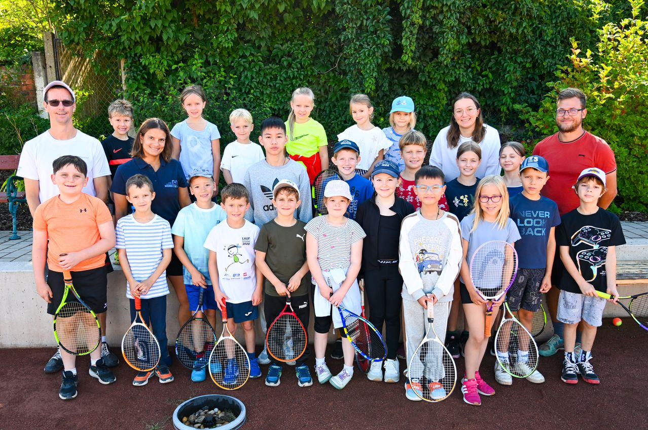 Eine Gruppe von Kindern und einer Frau steht zusammen und hält Tennisschläger, lächelt für ein Foto. Sie stehen vor einer Wand und einem grünen Busch.