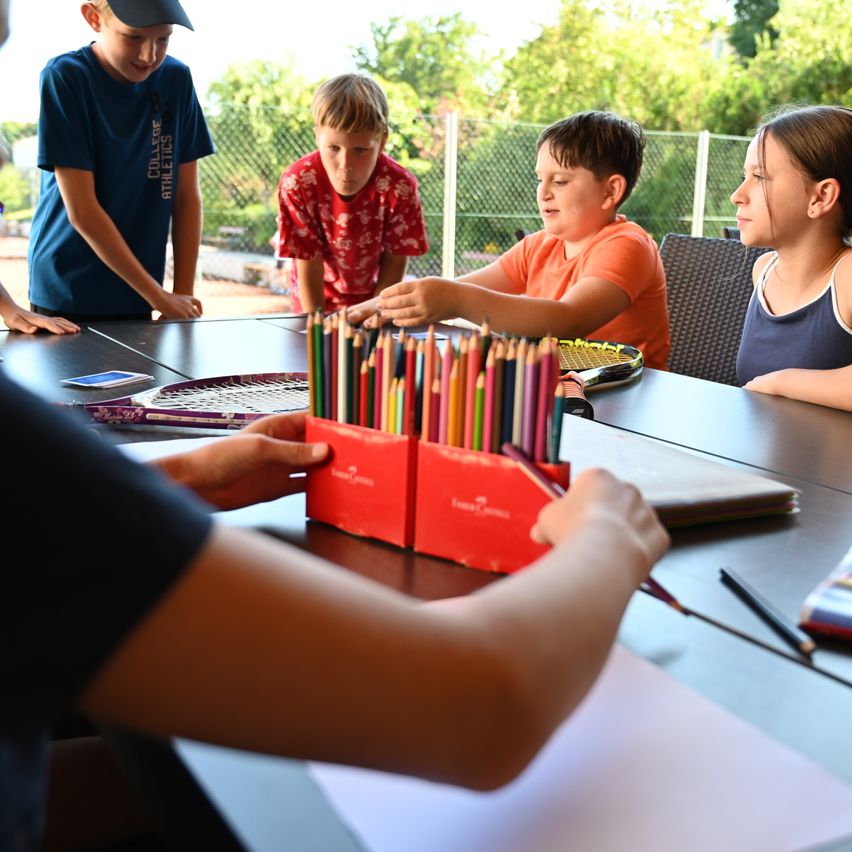 Eine Gruppe von Kindern sitzt an einem Tisch und ist mit farbigen Stiften und Papier beschäftigt. Ein Junge lehnt sich vor und ein anderer Junge sitzt in der Nähe, beide konzentrieren sich auf die Aktivität.