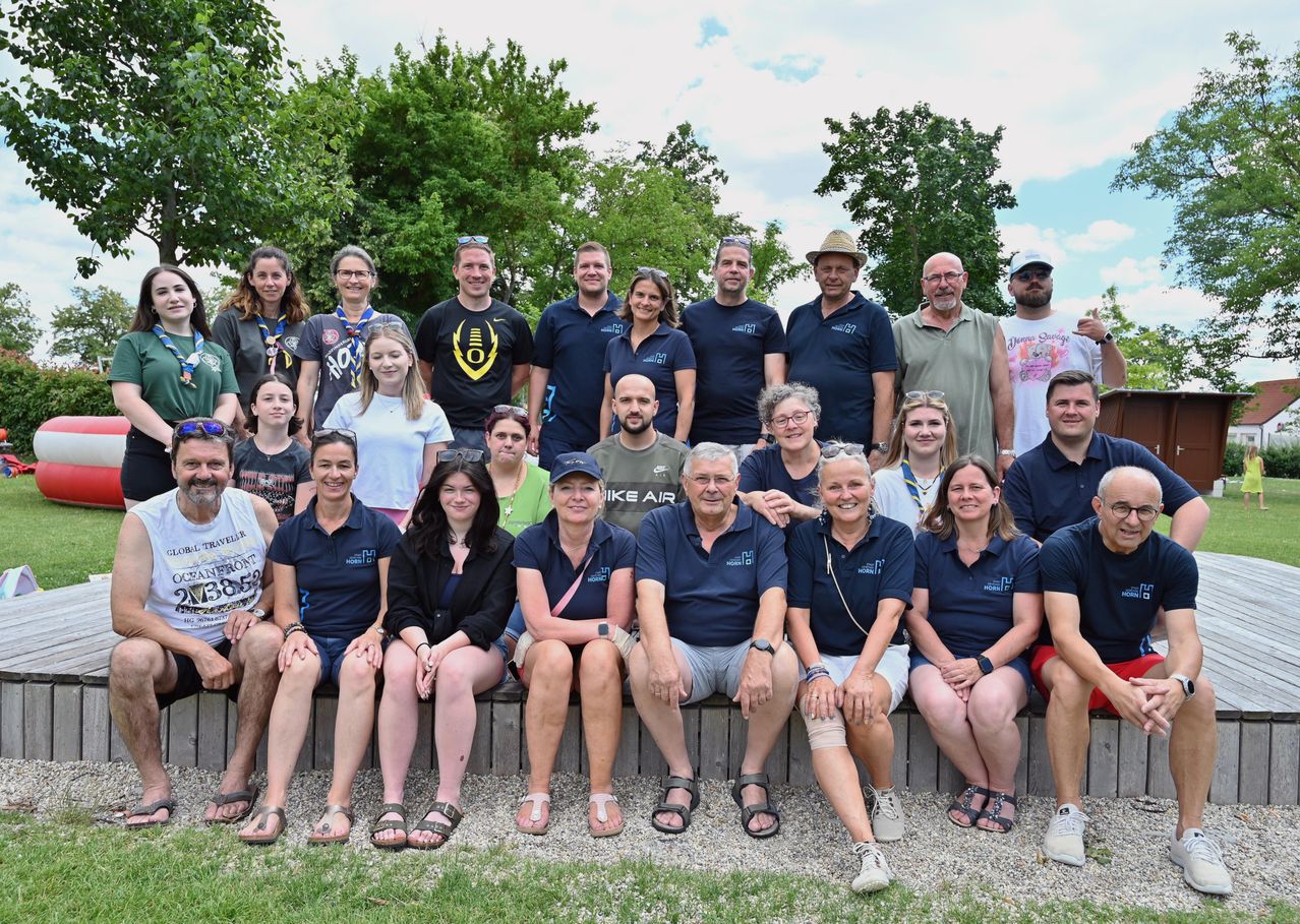 Eine Gruppe von Menschen in passenden blauen Poloshirts posiert für ein Foto im Freien in einem Park. Einige tragen Hüte, Brillen und Armbänder. Bäume und ein blauer Himmel sind im Hintergrund.
