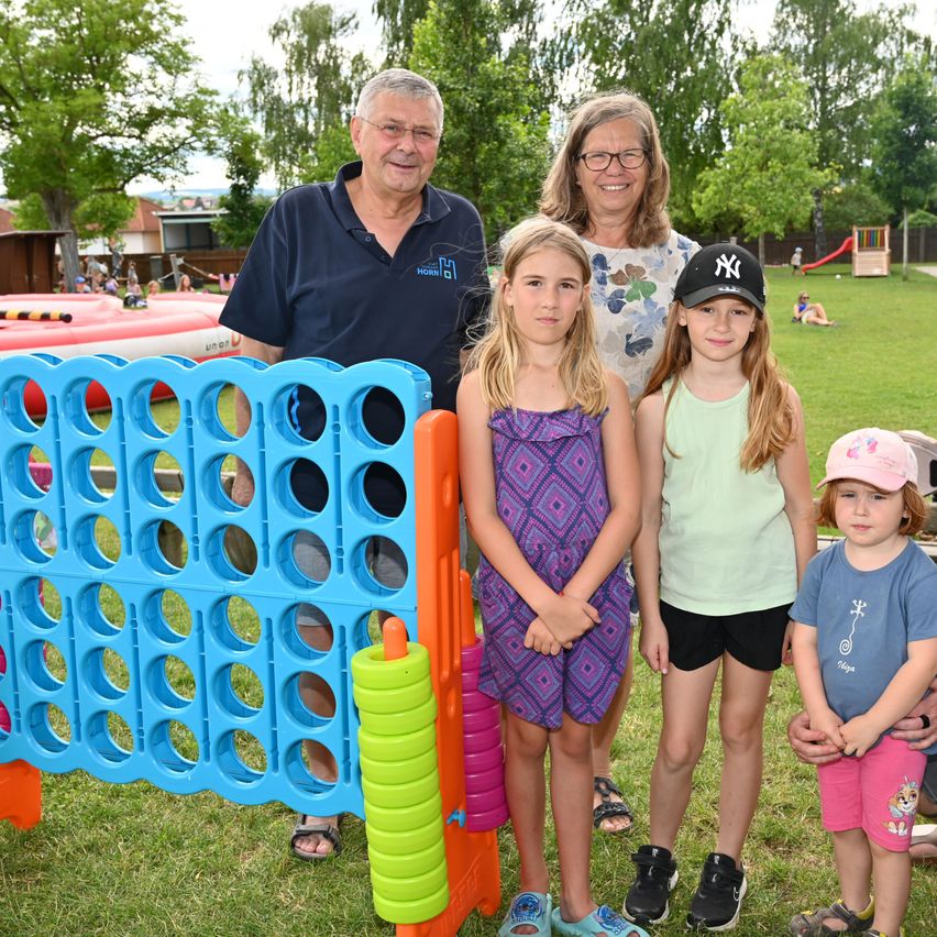Eine vierköpfige Familie steht vor einem großen, bunten Spielbrett und lächelt für die Kamera. Dahinter ist ein aufblasbares Spielgerät zu sehen, und in der Ferne sind weitere Spielgeräte zu erkennen.