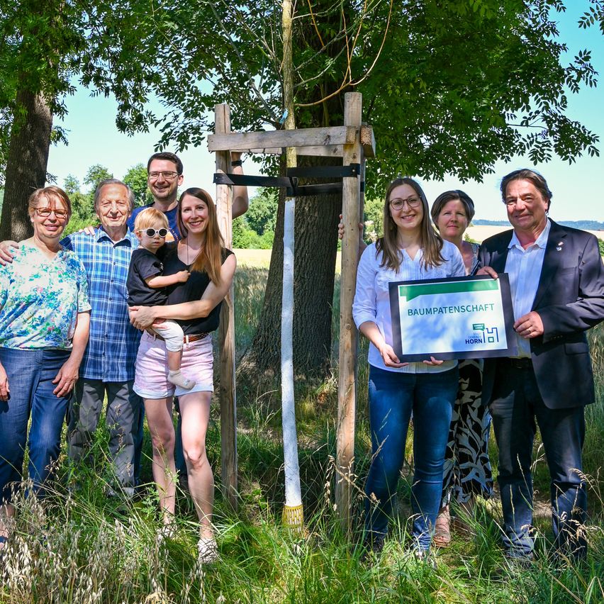 Eine Familie und einige Personen posieren für ein Foto auf einem Feld mit einem Baum und einem Schild, das Baumpatenschaft besagt. Die Familie besteht aus einer Frau, die ein Baby hält, einem Mann mit Brille und anderen Personen.