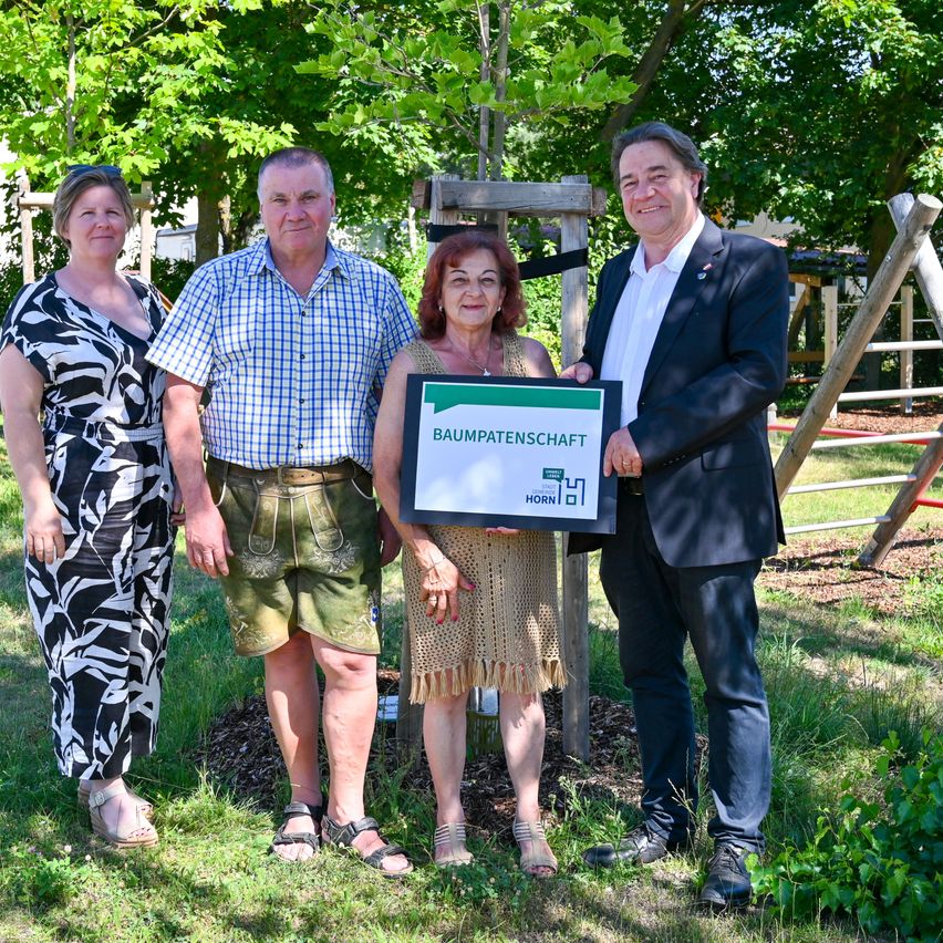 Vier Personen stehen in einem Park mit einem Schild in der Hand. Das Schild heißt Baumpatenschaft. Dahinter befindet sich eine Holzstruktur. Der Mann im Anzug hält das Schild mit beiden Händen.
