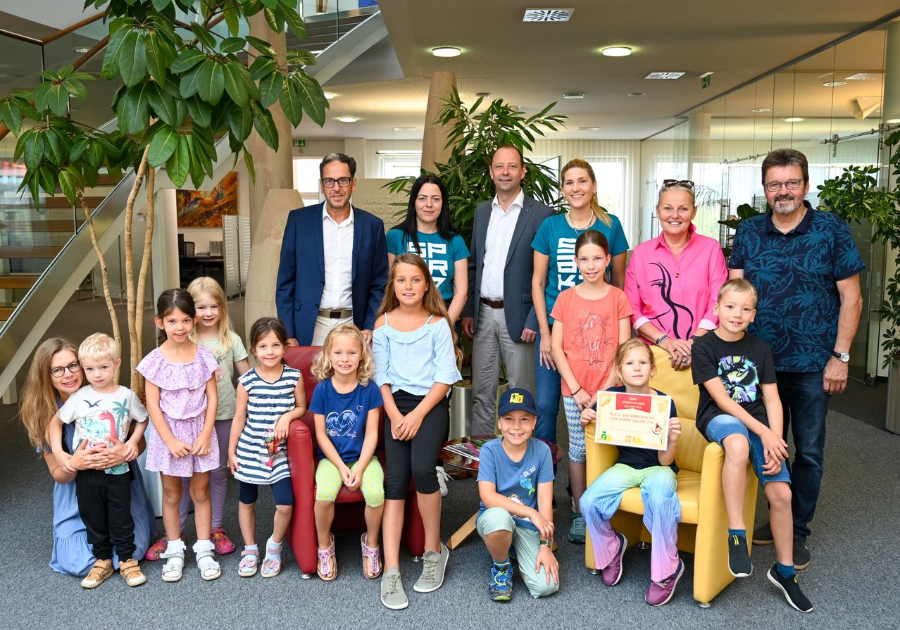 Eine Gruppe von Kindern und Erwachsenen posiert für ein Foto in einem modernen Büro mit großen Pflanzen und Glaswänden. Einige Kinder sitzen auf Stühlen, während andere hinter Erwachsenen stehen, einer hält ein Zertifikat.
