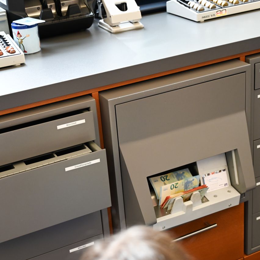 Ein grauer Bürotresen mit offenen Schubladen und einem Geldzählautomaten, der Schweizer Franken zeigt. Verschiedene Büromaterialien sind auf dem Tresen platziert.