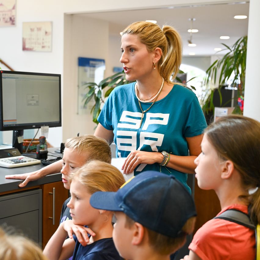 Eine Frau steht vor einer Gruppe von Kindern in einem Büro und hält eine Präsentation. Sie trägt ein blaues T-Shirt mit weißen Texten. Die Kinder hören aufmerksam zu, mit einem Computermonitor und einer Tastatur hinter ihnen.